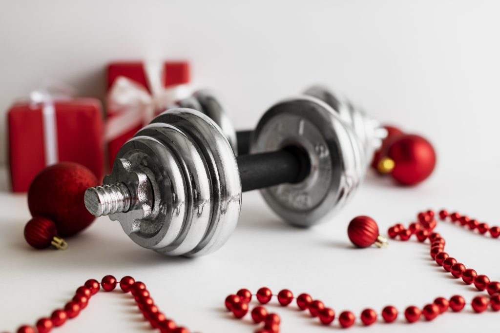 Chrome dumbbell surrounded by red Christmas ornaments and holiday decorations, symbolizing festive fitness and holiday workout motivation.